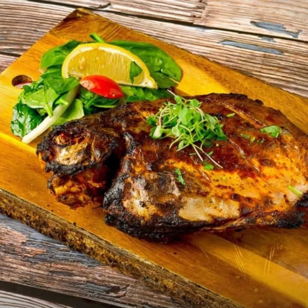 Grilled Indian fish, garnished with fresh greens, lemon, and tomato, served on a wooden board.