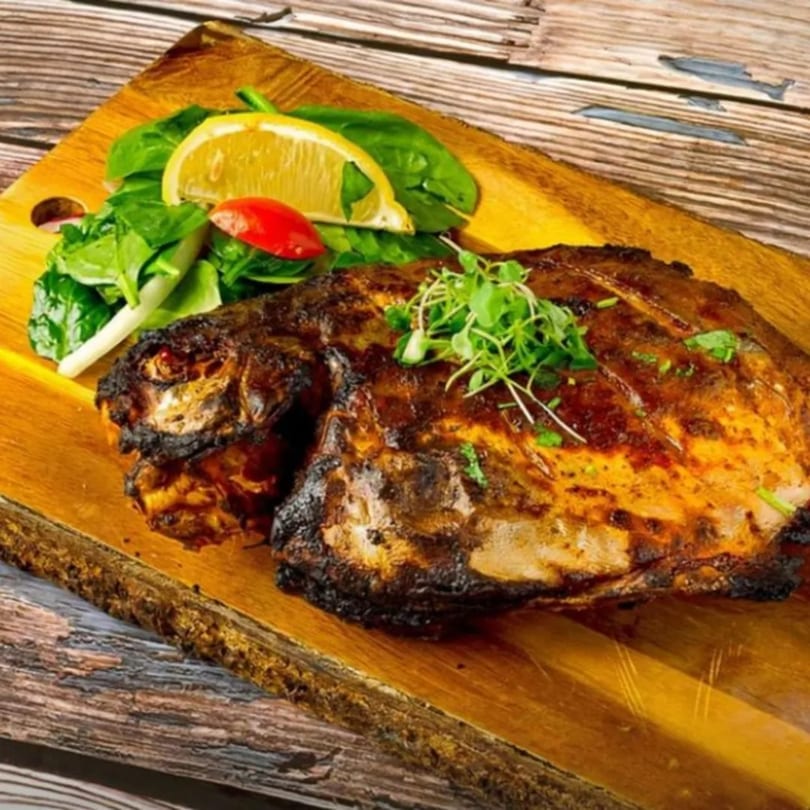 Grilled Indian fish, garnished with fresh greens, lemon, and tomato, served on a wooden board.