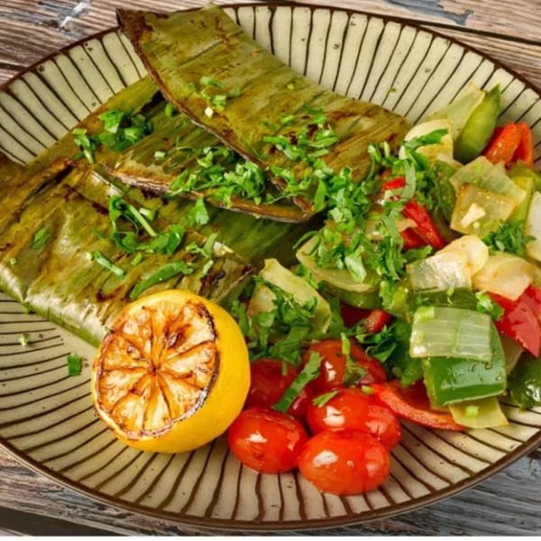 Indian dish with food wrapped in banana leaves, served with grilled lemon, tomatoes, and vegetables.